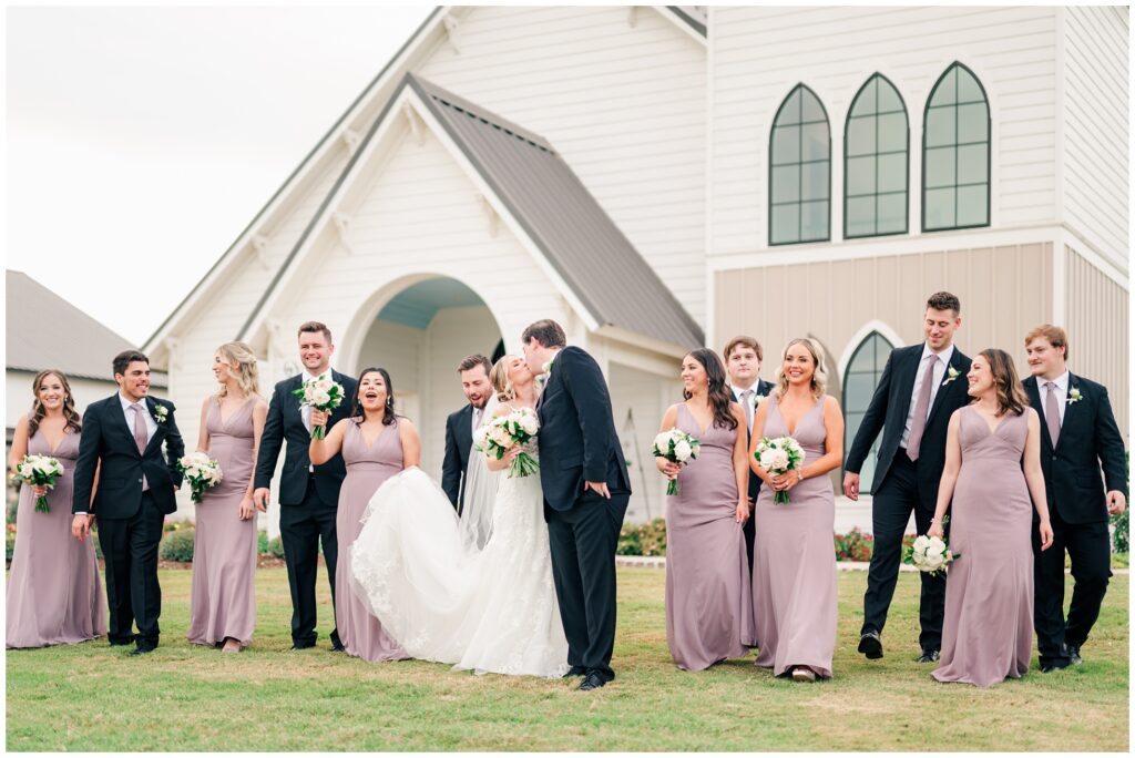 Wedding Party of Bridesmaids and Groomsmen surround the happy couple kissing in front of the deep in the heart farms wedding chapel.